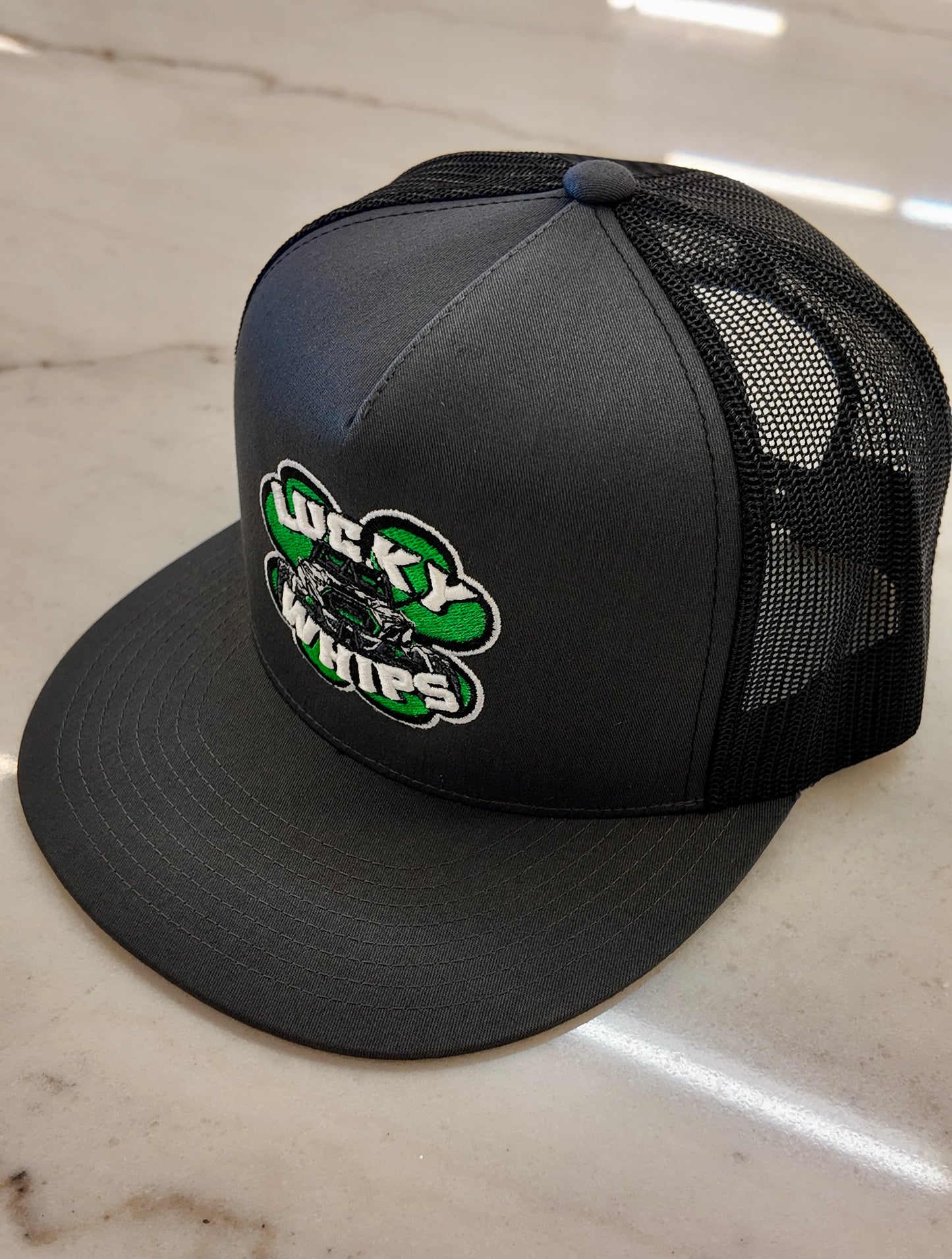 Charcoal trucker flat bill hat by Lucky Whips with mesh back and adjustable snap closure.