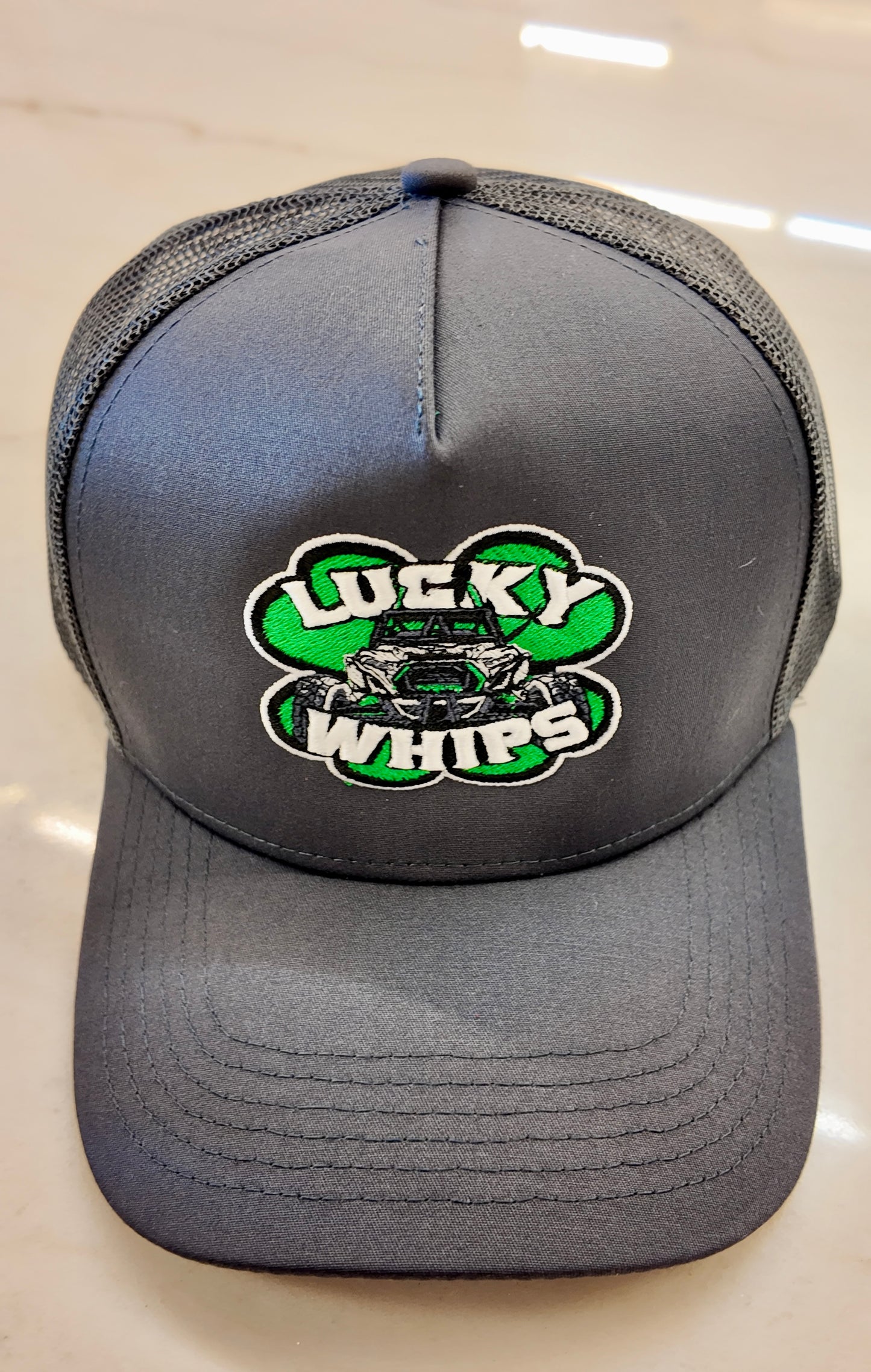 Charcoal trucker hat with a curved bill by Lucky Whips, featuring a breathable mesh back and adjustable snap closure.