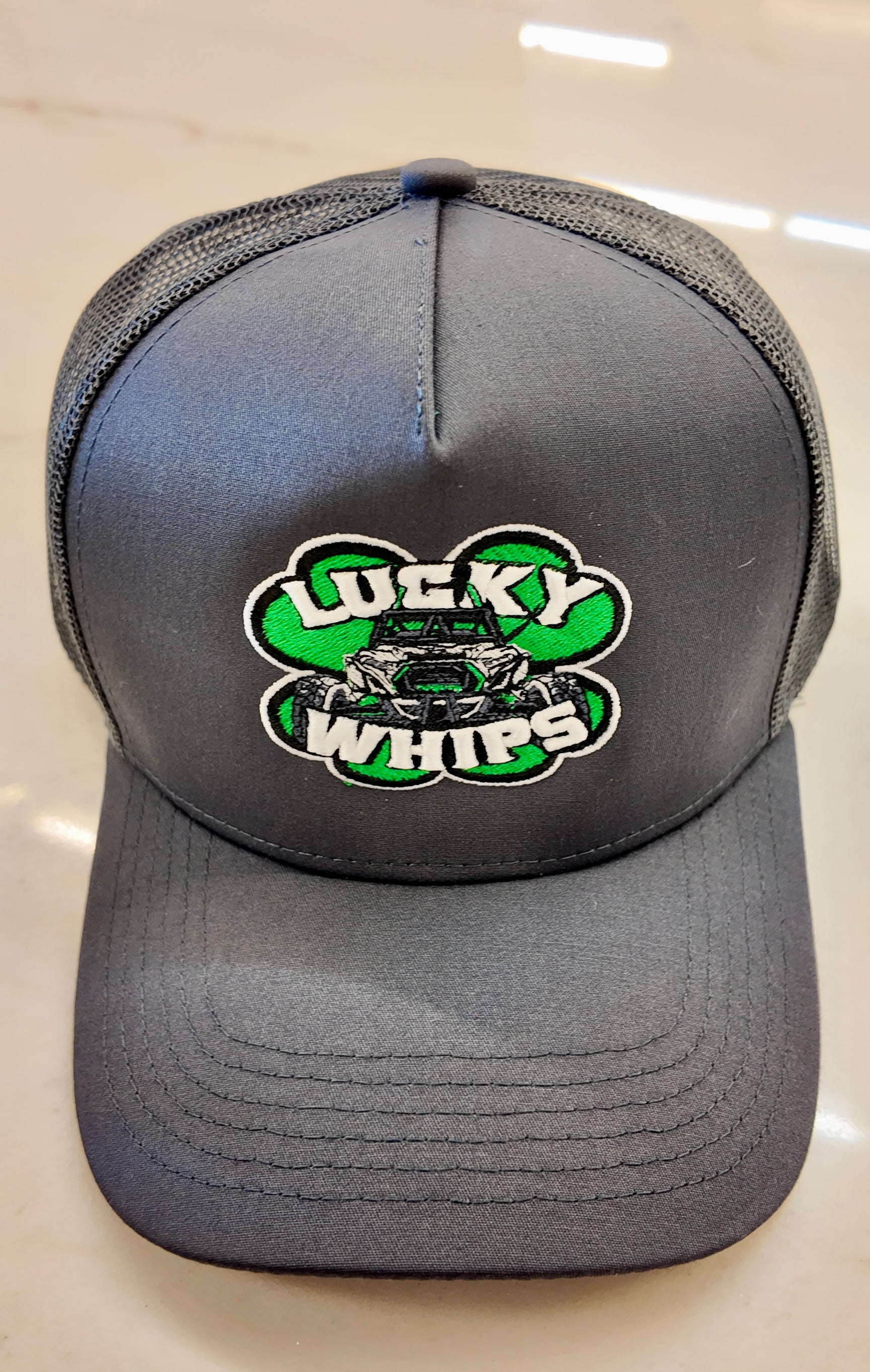 Charcoal trucker hat with a curved bill by Lucky Whips, featuring a breathable mesh back and adjustable snap closure.